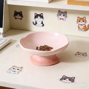 May include: A pink ceramic pet bowl with a scalloped edge, containing dry pet food. The bowl is on a pedestal base. Cat stickers are visible in the background.