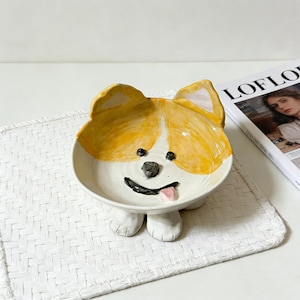 May include: A ceramic dog-shaped bowl with a yellow and white painted design. The bowl has a smiling face with a black nose, eyes, and mouth, and a pink tongue. The bowl is resting on two white paws.