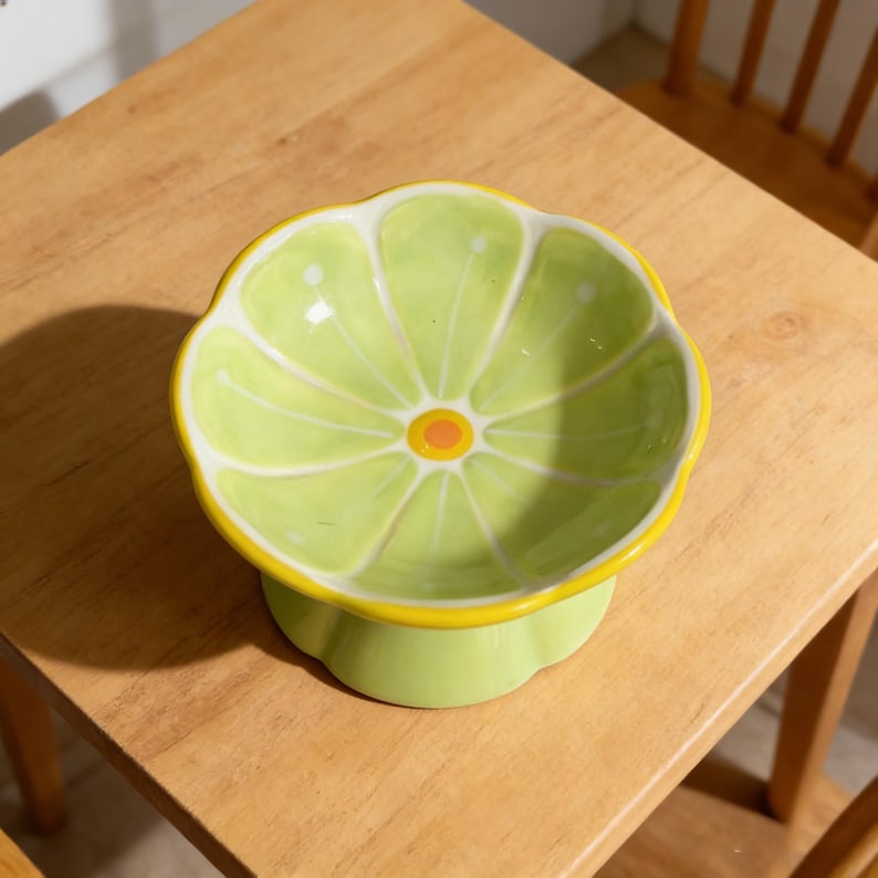Peut inclure: Un bol en c&eacute;ramique en forme de tranche d'agrume avec un int&eacute;rieur vert clair pr&eacute;sentant des segments radiaux blancs et un petit centre orange. Le bol poss&egrave;de un bord festonn&eacute;, un contour jaune vif et une base sur pied vert clair assortie.