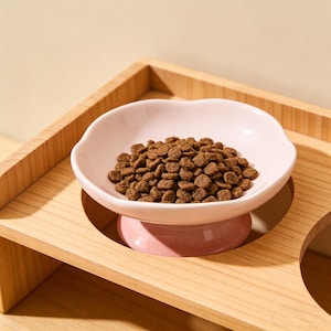May include: A pink, heart-shaped pet food bowl filled with brown kibble, set on a light pink pedestal. The bowl sits in a wooden frame with a natural finish, designed to hold the bowl securely. The image is well-lit, highlighting the bowl's shape.