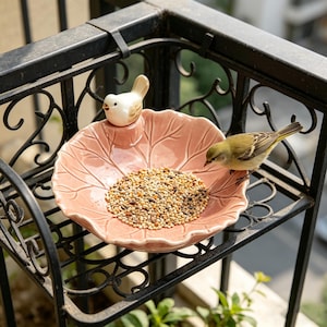 Puede incluir: Un comedero para pájaros de cerámica rosa con forma de hoja, lleno de alpiste. Un pequeño pájaro de cerámica se posa encima, y un pájaro vivo se posa en el borde. El comedero está montado en una barandilla de balcón de metal negro.