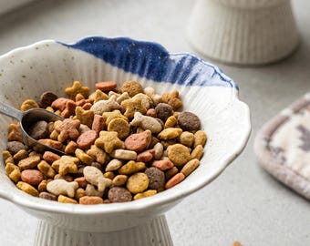 May include: A ceramic pedestal bowl with a white speckled finish and a blue-dipped scalloped rim, filled with assorted brown, tan, and orange dry pet food in star and fish shapes, accompanied by a small metal scoop resting inside the bowl.