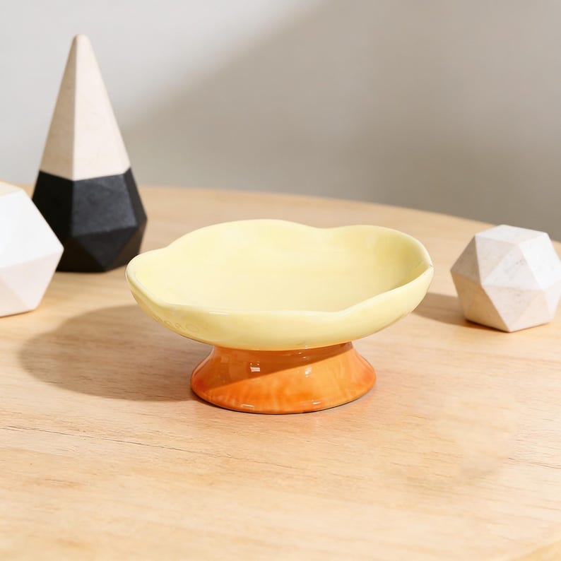 May include: A small, ceramic dish with a scalloped edge, in a pale yellow colour. The dish sits on a pedestal base that transitions from orange to yellow. Decorative wooden geometric shapes are in the background.