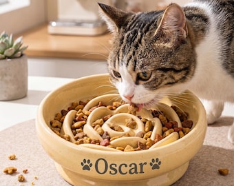 May include: A beige ceramic slow feeder bowl with a spiral interior design, filled with brown dry pet food. The exterior features the name Oscar printed in dark grey text, flanked by two paw print icons. A tabby cat is eating from the bowl.