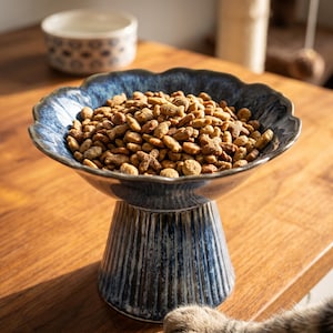 May include: A ceramic pet food bowl with a scalloped rim and a fluted pedestal base, featuring a reactive blue and white glaze finish. The bowl is filled with dry brown kibble in various shapes, including small fish and cross forms, for animal feeding.