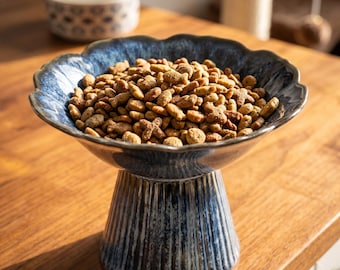 May include: A ceramic pet food bowl with a scalloped rim and a fluted pedestal base, featuring a reactive blue and white glaze finish. The bowl is filled with dry brown kibble in various shapes, including small fish and cross forms, for animal feeding.