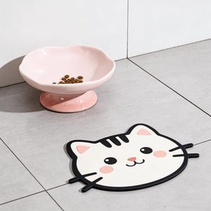 May include: A pink ceramic pet bowl with a scalloped edge, containing brown kibble, is on a pedestal. Next to it is a white cat-shaped placemat with black and pink details, including whiskers and rosy cheeks.