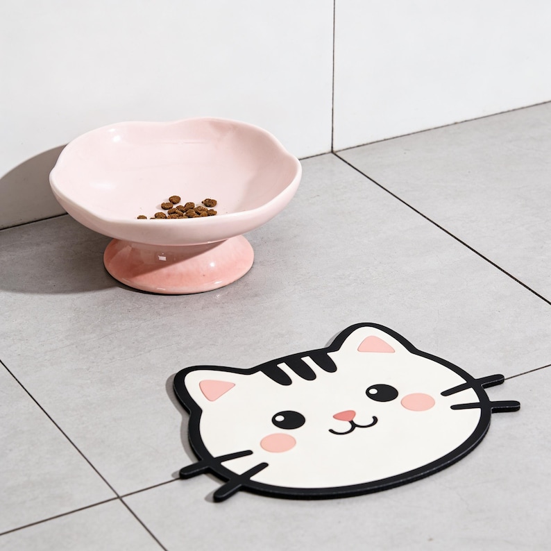 May include: A pink ceramic pet bowl with a scalloped edge, containing brown kibble, is on a pedestal. Next to it is a white cat-shaped placemat with black and pink details, including whiskers and rosy cheeks.