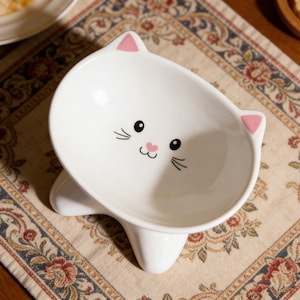 May include: A white ceramic bowl shaped like a cat head with two pointed ears featuring pink accents. The interior of the bowl displays a printed cat face with black eyes, black whiskers, and a small pink heart-shaped nose centered on the base.
