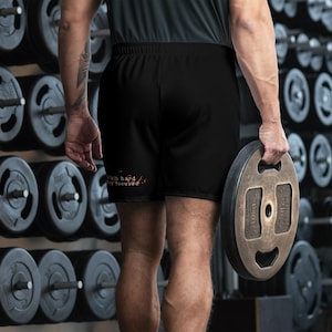 May include: Black athletic shorts with the text "Train hard, stay focused" in orange and brown. The shorts have an elastic waistband. A person is holding a 22-pound weight plate in a gym setting.