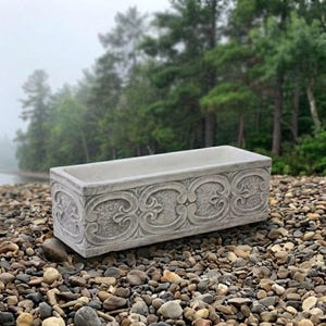 May include: A rectangular concrete planter with a light gray finish, featuring a repeating embossed scrollwork pattern along the front and side panels. The planter has a smooth top rim and a deep, open interior cavity for holding plants.