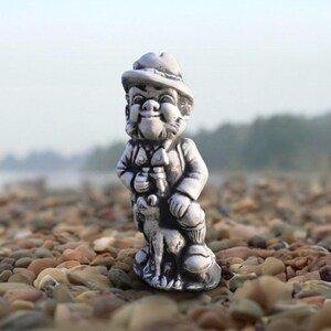 May include: A small, monochromatic gray figurine of a person wearing a wide-brimmed hat and a vest, standing next to a small dog. The figure has a detailed face with a prominent nose and is sculpted with a smooth, matte finish throughout the piece.