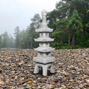 May include: A gray stone pagoda sculpture featuring a multi-tiered design with four levels, square openings on each tier, and a pointed finial top. The structure stands on four sturdy legs and displays a weathered, textured surface throughout.
