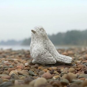 May include: A small, light gray bird figurine with a textured, stone-like finish. The bird is depicted in a resting position with its head turned slightly to the side, showing detailed carved feathers on its wings and a smooth, rounded body shape.