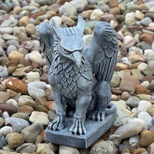 May include: A gray griffin statue with detailed feather and wing textures. The mythical creature has a bird-like head with a sharp beak and large, spread wings. The statue is set on a rectangular base and is surrounded by small rocks.