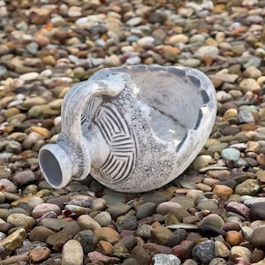 May include: A weathered, light gray ceramic vessel with a rounded body and a single side handle. The surface features a textured, mottled finish with a geometric, carved pattern of concentric lines and a wide, open top with a jagged, uneven rim.