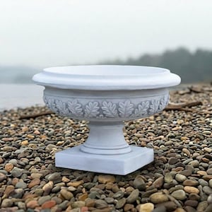 May include: A white stone planter with a wide, circular basin and a thick, rounded rim. The exterior of the bowl features a repeating relief pattern of stylized oak leaves. The planter rests on a pedestal base with a square bottom.