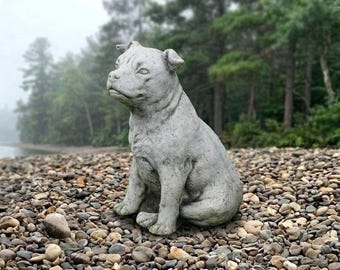 May include: A gray, stone-textured sculpture of a seated dog with pointed ears, a broad snout, and a muscular build. The statue is depicted in a sitting position with its front paws planted firmly on the ground and its head turned slightly to the left.