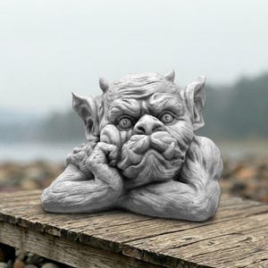 May include: A grey stone-colored gargoyle sculpture featuring a creature with pointed ears, horns, and a furrowed brow. The figure rests its chin on one hand, displaying detailed facial features, including wide eyes and prominent tusks.