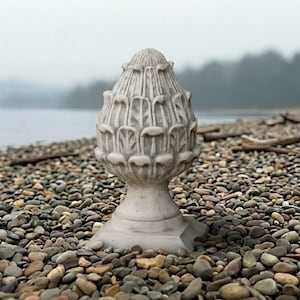 May include: A light gray stone finial featuring a carved, stylized pinecone or artichoke design. The object has a rounded, textured top with repeating leaf-like patterns and sits on a smooth, flared pedestal base with a square bottom.