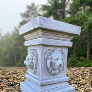 May include: A square light gray stone pedestal featuring a relief carving of a lion head on each visible side. The structure has a wide flat top, a tapered middle section with recessed panels for the lion motifs, and a sturdy base with tiered edges.