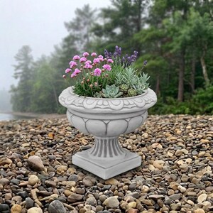 May include: A light gray stone-look planter with a fluted pedestal base and a decorative scalloped rim. The planter is filled with a variety of small green succulents and clusters of small pink and purple flowers arranged in a rounded display.
