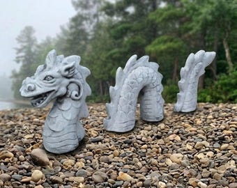 Puede incluir: Una escultura de dragón de piedra gris que consta de tres piezas separadas: una cabeza detallada con un cuerno curvado, una sección central arqueada con escamas texturizadas y un segmento de cola puntiagudo, dispuestos como si emergieran del suelo.