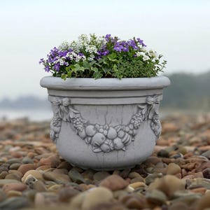 May include: A light gray stone planter with a wide rim and a relief carving of fruit swags and bows around the exterior. The planter is filled with a dense arrangement of small white and purple flowers mixed with green foliage.