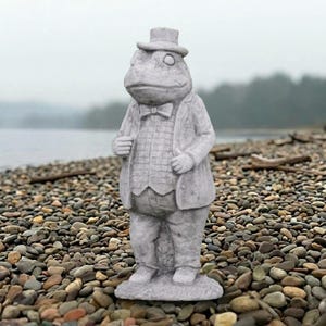 May include: A gray stone statue of a standing frog dressed in a formal suit, waistcoat, bowtie, and top hat. The frog has a rounded belly and is depicted in a human-like posture with hands resting near the waist, featuring a textured stone finish.