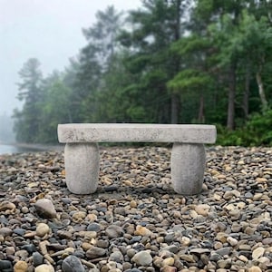 May include: A light gray stone bench featuring a long, rectangular seat supported by two thick, rounded, pillar-like legs. The bench has a textured, weathered surface and is positioned on a ground covered in various small, smooth, rounded stones.