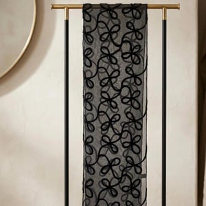 May include: A rectangular black sheer scarf featuring a continuous embroidered pattern of interconnected loops. The scarf is draped over a gold and black metal display stand with a wooden base, showcasing the intricate black thread work.