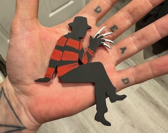 May include: A black and red paper cutout of a seated figure with a hat and bladed glove. The figure is wearing a red and black striped shirt and black pants. The figure is a silhouette of a horror movie character.