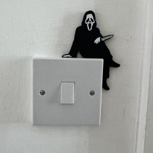 May include: A black and white Ghostface figure from the Scream films, perched on a white light switch. The figure is holding a knife. The light switch is mounted on a white wall. A quirky home decor item.