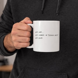 Peut inclure: Mug en céramique blanche avec le texte "git add . git commit -m "please work" git push" en noir. La tasse a une anse blanche et est tenue par une personne.