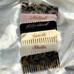 May include: A stack of personalized combs in various colors, including tortoiseshell, pink, black, cream, and white. Each comb features a name or initials in gold lettering. The combs are arranged on a white, textured surface.