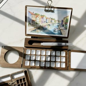 May include: A dark wood watercolor palette box featuring twenty-four empty white paint pans, a central slot for a water brush, and a hinged lid with a metal clip holding a landscape painting of a canal with colorful buildings and boats.