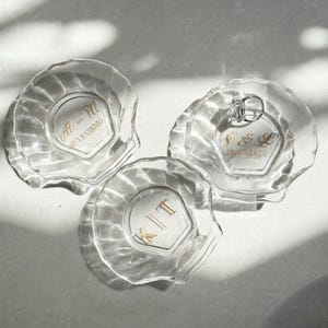 May include: Three clear glass shell-shaped trinket dishes with scalloped edges. Each dish features gold-colored custom lettering and dates. One dish displays the initials M + M with a date, another K | T, and the third P & L with a date and a silver ring.