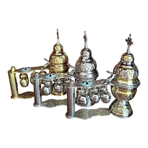 May include: Two ornate metal incense burners featuring a tiered, dome-shaped top with a cross finial, intricate floral engravings, and a handle attached to a base with small hanging bells and a circular blue and white decorative accent piece.