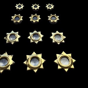 Puede incluir: Una colección de objetos decorativos dorados en forma de estrella. Cada pieza presenta un elemento central circular rodeado por un diseño de estrella. Los objetos están dispuestos sobre un fondo negro.