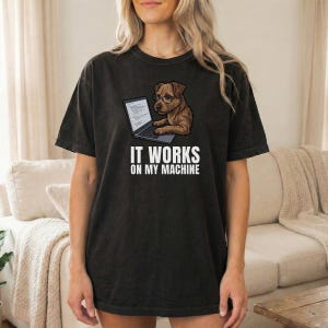 Op de afbeelding: Zwart t-shirt met een afbeelding van een bruine hond met een bril die op een laptop werkt. De tekst "IT WORKS ON MY MACHINE" staat eronder. Het shirt is donker van kleur en heeft een losse pasvorm.