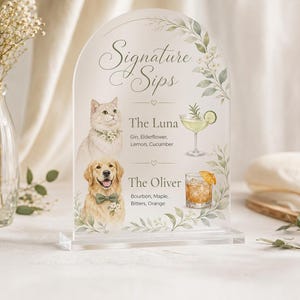 May include: An arched acrylic sign titled Signature Sips featuring illustrations of a cat and a golden retriever. The text lists The Luna with gin, elderflower, lemon, cucumber and The Oliver with bourbon, maple, bitters, orange, framed by green leaves.