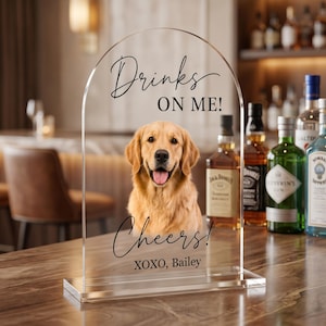 Personalized acrylic pet wedding bar sign featuring a golden retriever portrait with “Drinks On Me” wording, elegant script text, and custom pet name detail. This custom dog signature drink sign is perfect for wedding bar decor, cocktail hour, reception table styling, bridal shower, engagement party, or pet-themed celebration. Available in clear, white, frosted, and black acrylic, it adds a charming and playful touch to your event setup.
