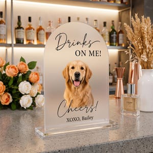 May include: An arched, frosted acrylic sign featuring a portrait of a golden retriever. The text reads "Drinks ON ME!", "Cheers!", and "XOXO, Bailey" in black script and sans-serif fonts. The sign is mounted on a clear rectangular base.