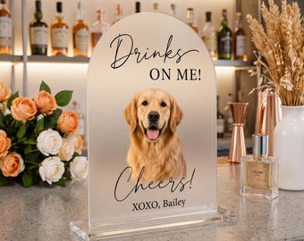 Op de afbeelding: Een boogvormig, mat acrylbord met een portret van een golden retriever. De tekst "Drinks ON ME!", "Cheers!" en "XOXO, Bailey" staat in zwarte letters op het bord. Het bord is bevestigd op een doorzichtige rechthoekige voet.