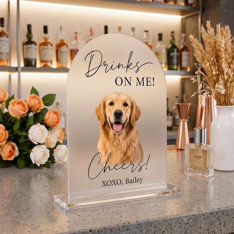 Personalized acrylic pet wedding bar sign featuring a golden retriever portrait with “Drinks On Me” wording, elegant script text, and custom pet name detail. This custom dog signature drink sign is perfect for wedding bar decor, cocktail hour, reception table styling, bridal shower, engagement party, or pet-themed celebration. Available in clear, white, frosted, and black acrylic, it adds a charming and playful touch to your event setup.