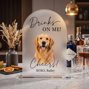 May include: An arched frosted acrylic sign featuring a portrait of a golden retriever. The text reads "Drinks ON ME!", "Cheers!", and "XOXO, Bailey" in black script and sans-serif fonts. The sign is mounted on a clear rectangular base.