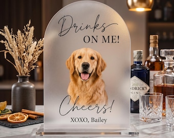 Op de afbeelding: Een boogvormig bord van mat acryl met een portret van een golden retriever. De tekst "Drinks ON ME!", "Cheers!" en "XOXO, Bailey" staat in zwarte letters op het bord. Het bord is bevestigd op een doorzichtige rechthoekige voet.