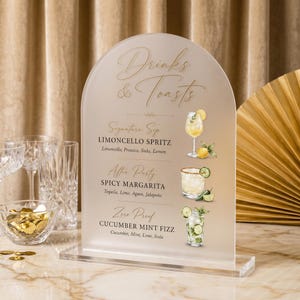 Può includere: Un cartello in acrilico smerigliato a forma di arco con il testo dorato Drinks & Toasts, che elenca tre cocktail: Limoncello Spritz, Spicy Margarita e Cucumber Mint Fizz. Ogni nome è accompagnato da un'illustrazione ad acquerello del drink.