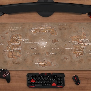 May include: A large, rectangular gaming mousepad featuring a detailed map of Azeroth with text. The pad is surrounded by gaming accessories: a black keyboard with red accents, a mouse, a controller, and a headset. The setup is on a wooden surface.