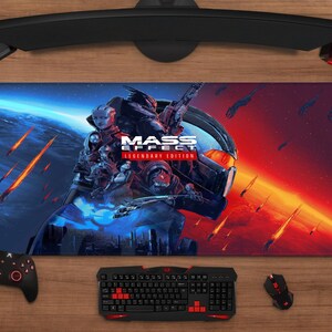 Pode incluir: Tapete de rato retangular para jogos com o logotipo e a arte de "Mass Effect: Legendary Edition". O design apresenta personagens e uma nave espacial contra fundos azuis e vermelhos. Um teclado, rato e controlador pretos também são visíveis.